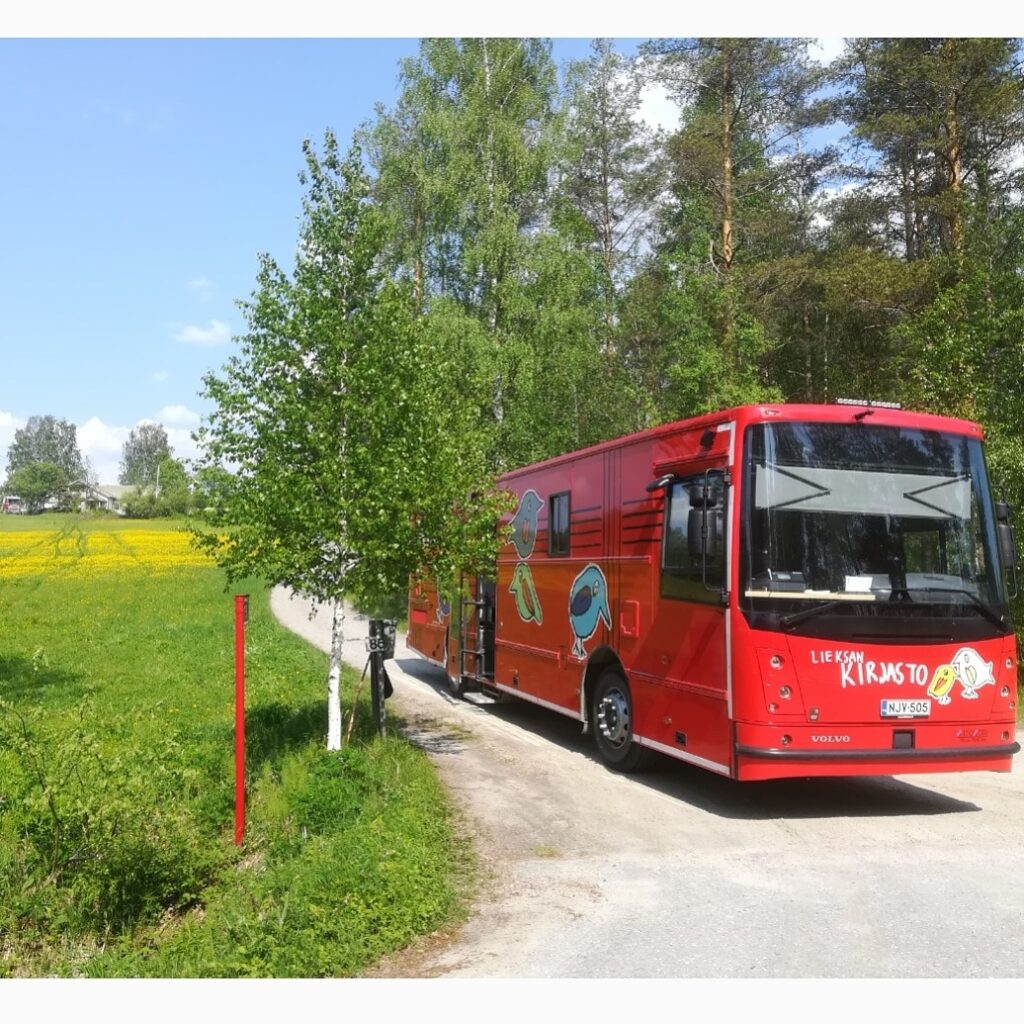 Kirjastoauto pysäköitynä tien risteykseen. Ympärillä näkyy puita ja kukkia täynnä oleva pelto.