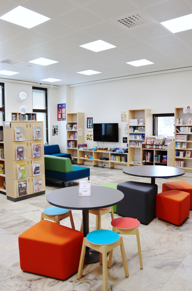 Lieksan kirjaston lasten- ja nuortenosastoa. Etualalla on kaksi pöytäryhmää. Taaempana on kirja- ja pelihyllyjä ja televisio.