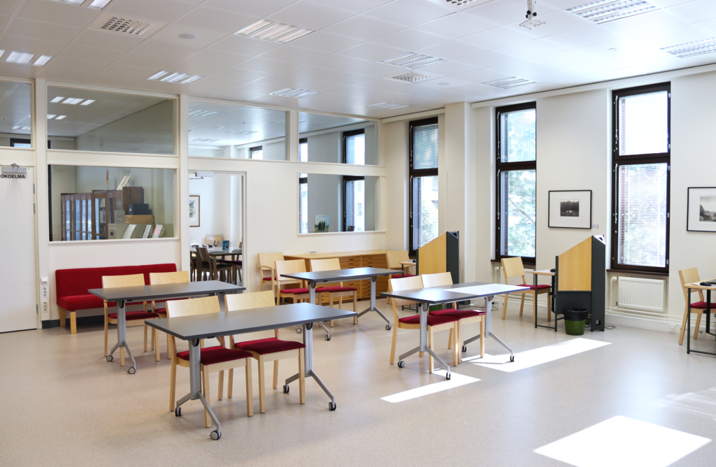Lieksan kirjaston matineatila. Tilassa on neljä pöytää ja tuoleja. Taustalla näkyvät kirjaston kotiseutukokoelmahuone ja mummolahuone.