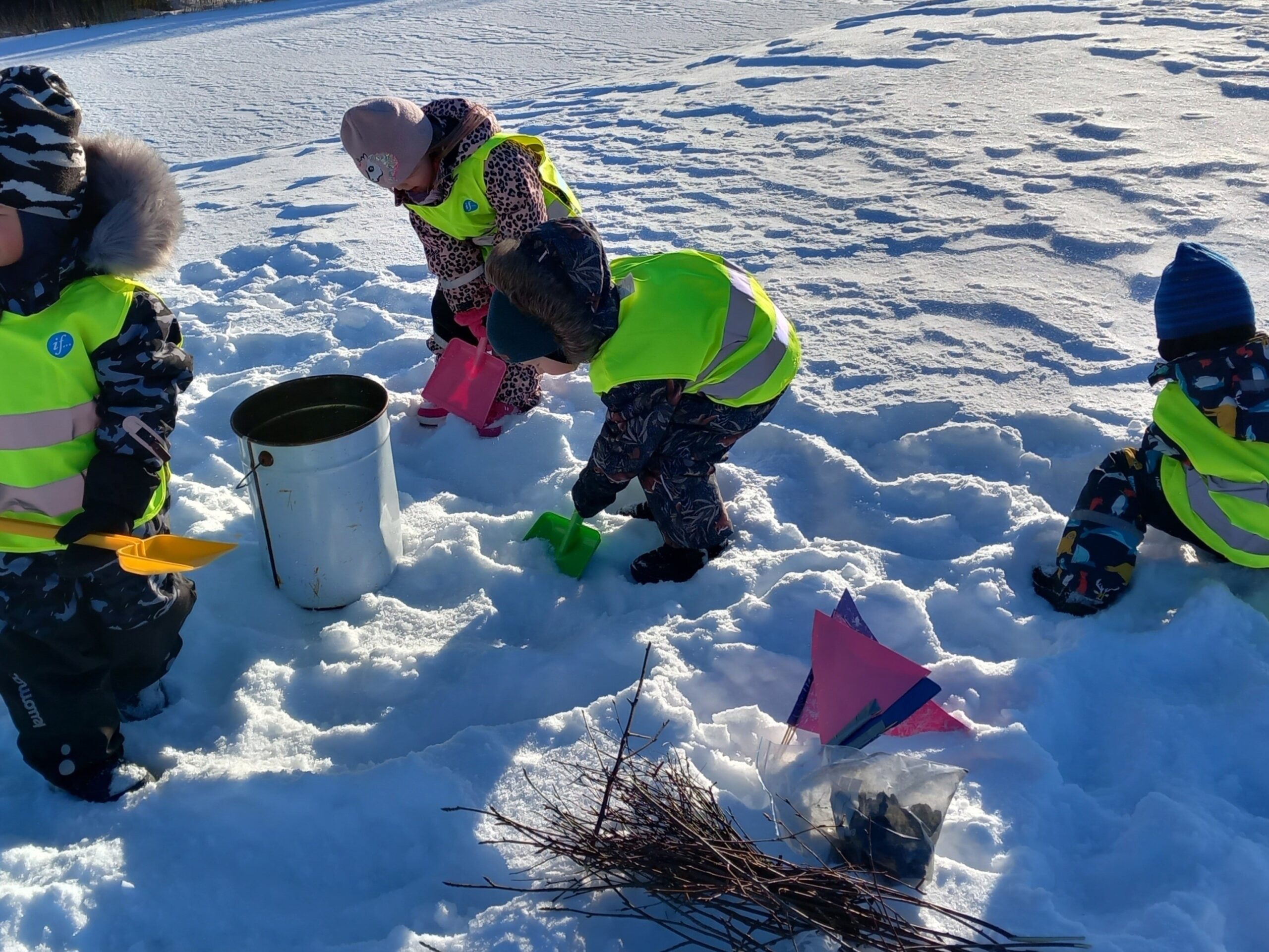 Lapsia lapioimassa lunta sankoon. Sankon vieressä kasa risuja.