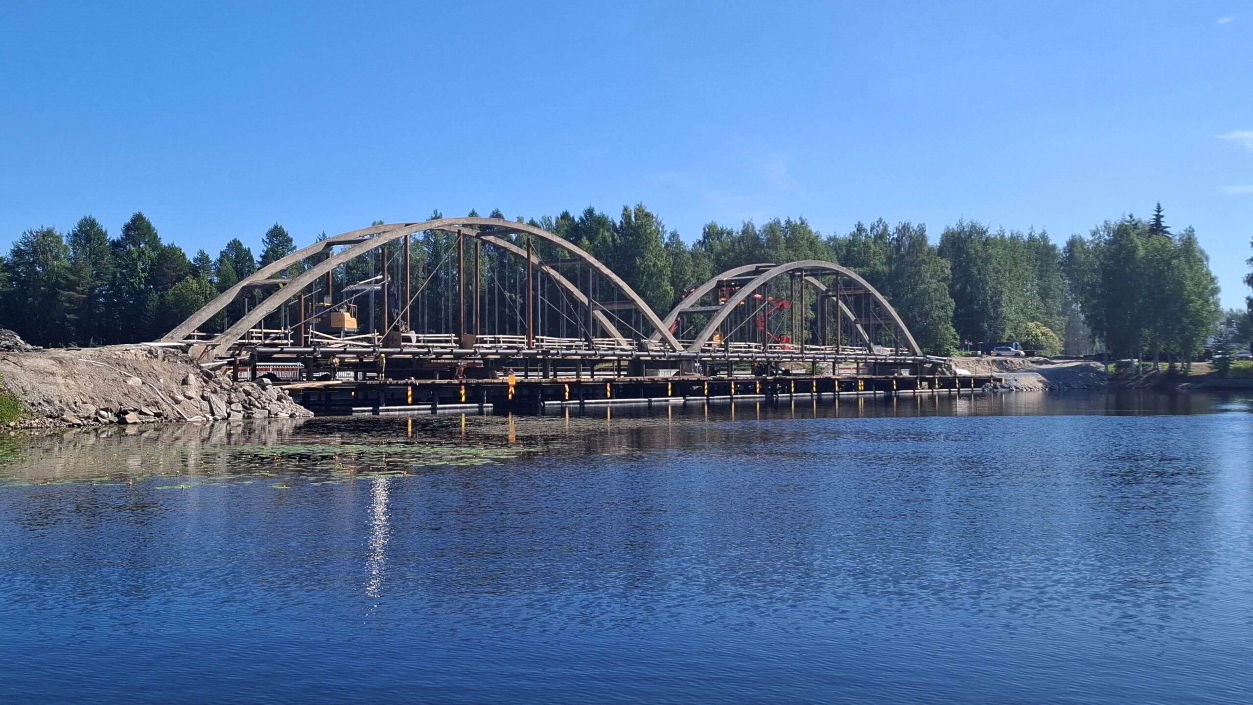 Kaksikaarinen silta. Sillalla työkoneita, vastarannalla auto ja koivuja.