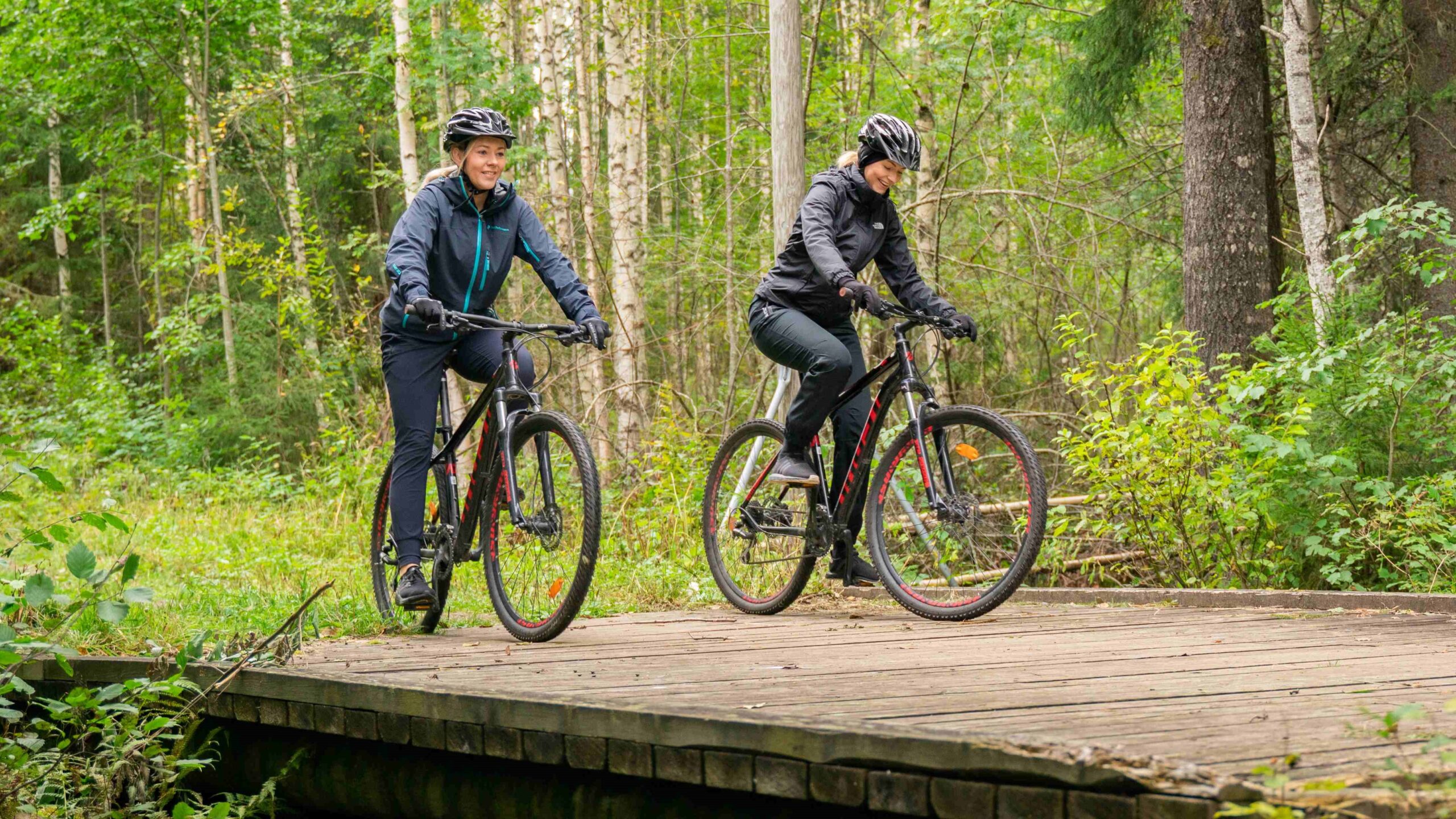 Kaksi naista metsässä ylittämässä siltaa maastopyörillä.