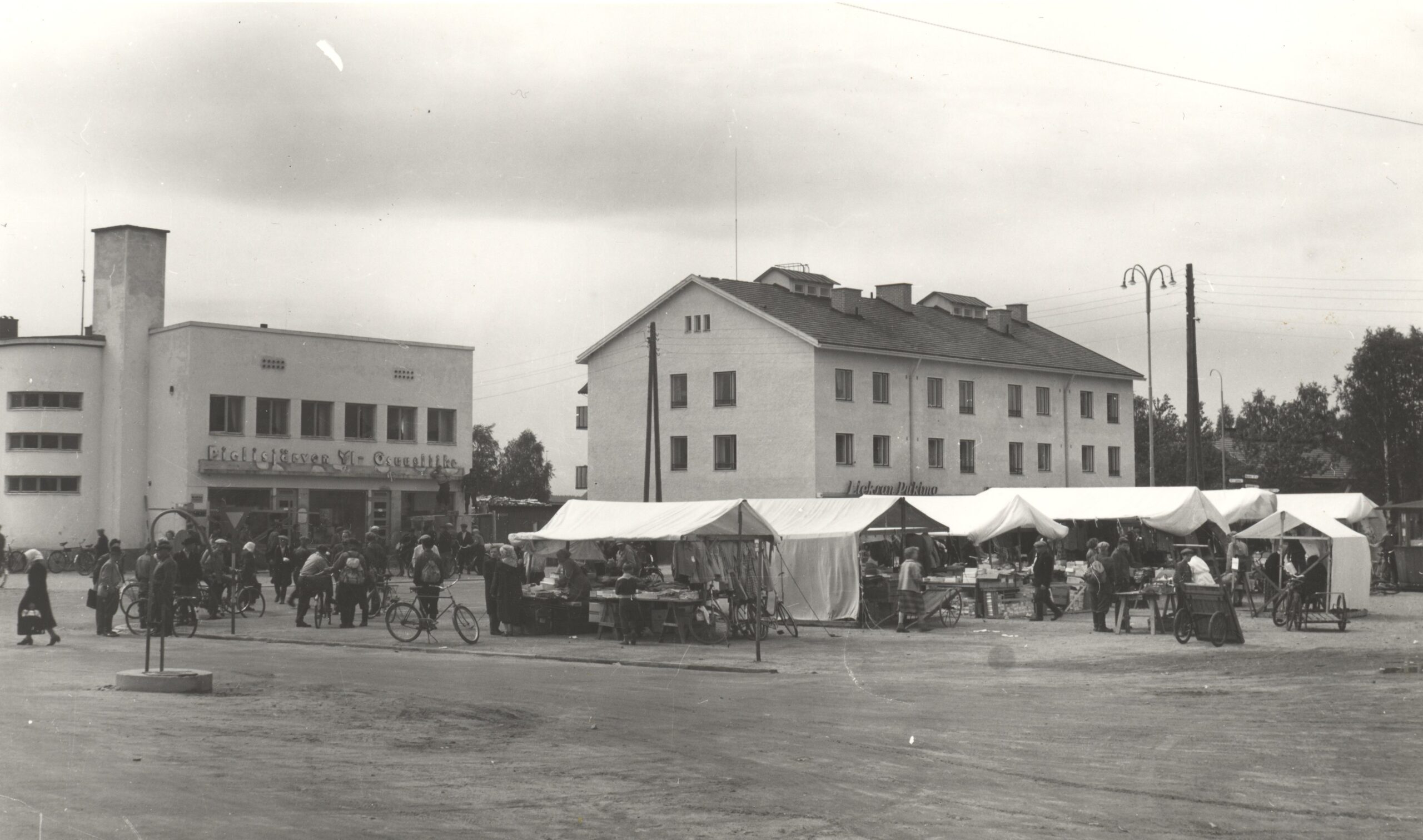 Vanha LIeksan tori Pielisentien varrella. Ihmisiä torikaupoilla myyntiktelttoja. Taustalla kaksi isoa rakennusta.